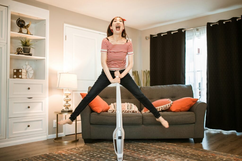 A joyful woman making cleaning fun by jumping with a vacuum in her living room.